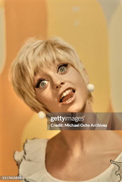 American actress Goldie Hawn, wearing a white scoop neck outfit with black trim, and white ball earrings, in a studio portrait, against a yellow...