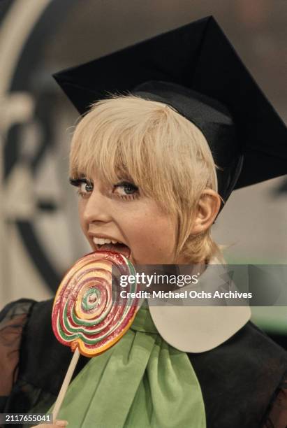 American actress Goldie Hawn, wearing a black outfit with a white collar, a green scarf and a mortarboard, holding a large multi-coloured lollipop,...