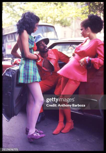 View of a trio of unidentified fashion models, one seated in the open door of a Cadillac Eldorado parked on an unspecified street, New York, New...