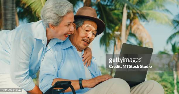 asian elderly man playing on laptop while sitting on a beach chair enjoys the sea view with his wife. a happy senior adult couple enjoys the travel lifestyle after retirement. - mes stock pictures, royalty-free photos & images