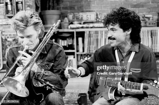 View of American Pop musicians Daryl Hall , on trombone, and John Oates, with an electric guitar, both of the duo Hall & Oates, as they sit on a low...