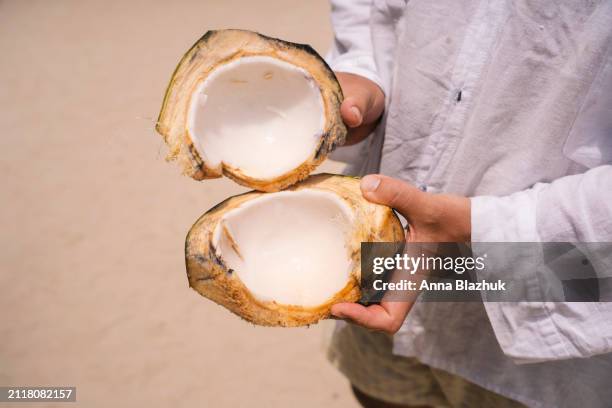 close-up of halved fresh coconut in hands of man on sand beach - coconut palm tree stock pictures, royalty-free photos & images