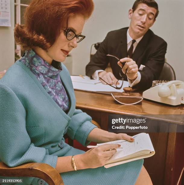 Office scene of a female secretary, wearing a pale blue wool skirt suit and patterned silk blouse, seated at a chair to take shorthand notes as,...