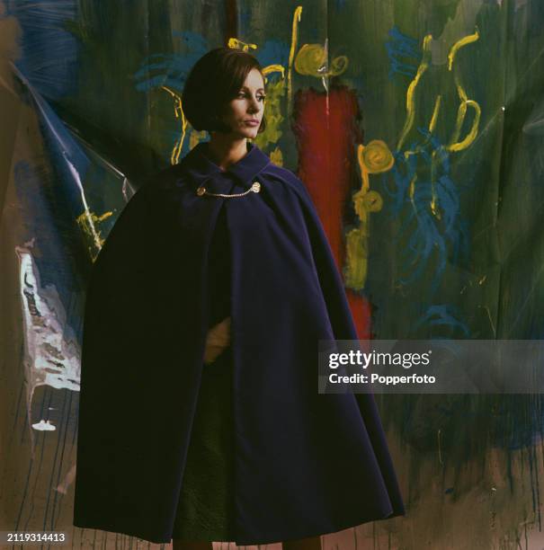Posed studio portrait of a female fashion model wearing a purple velvet cape over a green dress, she stands in front of a painted backdrop, London,...