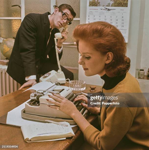 Office scene of a female secretary, wearing a light brown collarless jacket, seated at a desk to type up notes on her typewriter as, behind, a male...