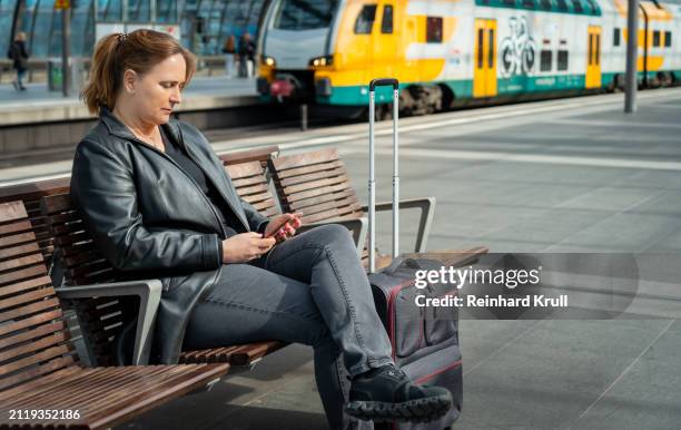 women sitting on a bench at railroad station and working with smartphone - reinhard krull stock pictures, royalty-free photos & images