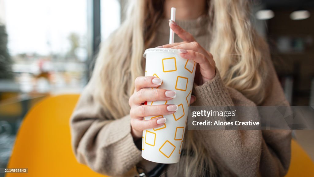 Beautiful girl in a beige sweater sits in a cafe and holds a paper cup of cola, close-up. Female hands with pretty nails holding cup of coffee