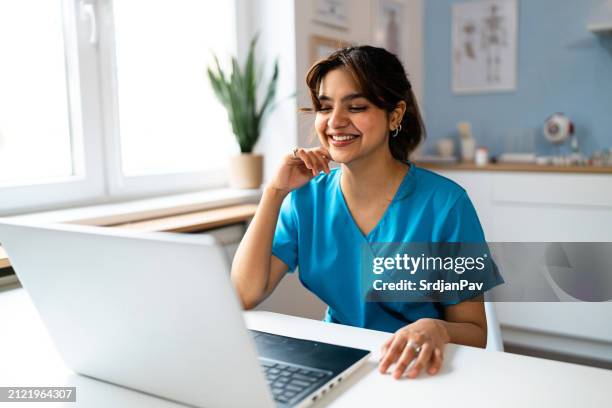 medizinstudentin indischer abstammung in blauen medizinischen kitteln, mit einem laptop bei der modernen ordination - medizinerkleidung stock-fotos und bilder