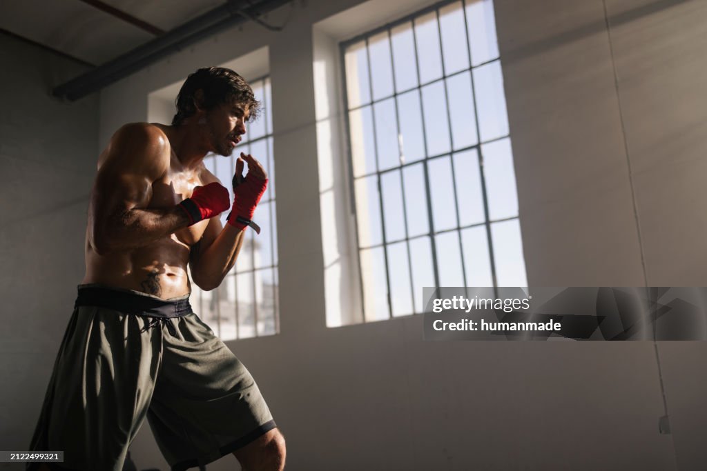 A focused male boxer with hand wraps preparing for a sparring session in a gym, illuminated by natural light from a window.