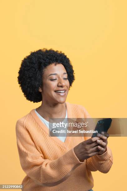 side view portrait of young black woman with afro hair smiling while using her smartphone on yellow background. - fashion suit stock pictures, royalty-free photos & images