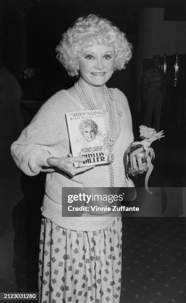 American comedian and actress Phyllis Diller, wearing a sweater and a skirt with square motifs, and a multi-strand beaded necklace, holding a copy of...