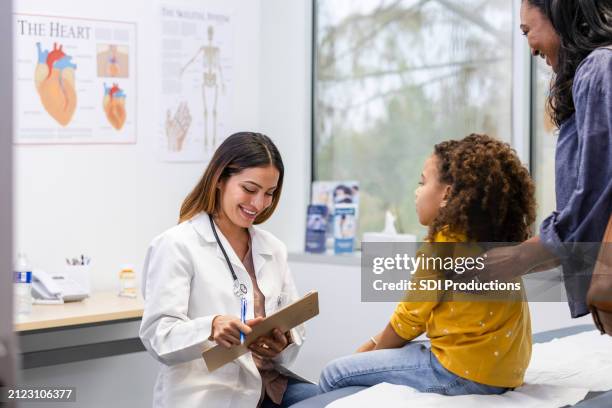 mid adult female doctor smiles while making notes in her patient's chart - cuidados de saúde primários imagens e fotografias de stock