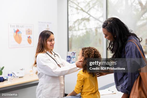 une mère se tient derrière sa fille pendant qu’elle est assise sur la table d’examen - blouse dexamen médical photos et images de collection