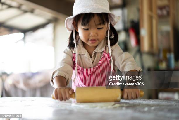 a focused little girl with a rolling pin works on flattening dough in a cooking class, wearing a cute bucket hat and apron. - asian pin up girls stockfoto's en -beelden