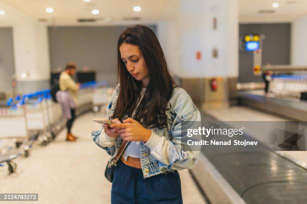 mulher jovem bonita usando seu smartphone enquanto espera por sua bagagem - área de retirada de bagagem - fotografias e filmes do acervo