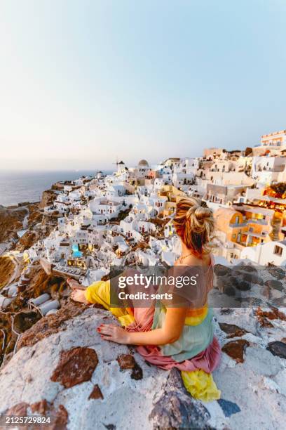 young woman enjoys traveling and looking at sunset view in front of beautiful summer vacation landscape of iconic blue domed churches along the caldera edge a sun setting against clear blue sky in oia, santorini, caldera, greek islands, greece - mediterranean-blue-roof-santorini stock pictures, royalty-free photos & images