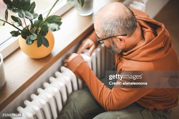 mature adult man warms himself by the radiator in the room - climate grief stock pictures, royalty-free photos & images