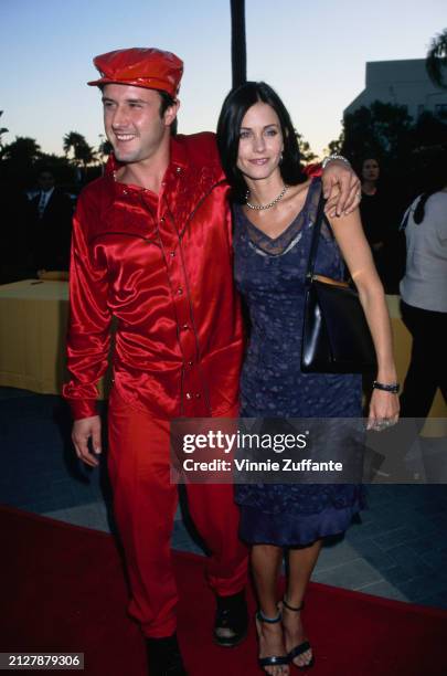 American actor David Arquette, wearing a red shirt with red trousers and a red peaked cap, with his arm around American actress Courteney Cox, who...