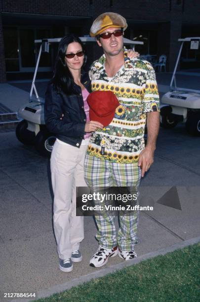 American actress Courteney Cox, wearing a denim jacket over a hooped pink top and white trousers, and her husband, American actor David Arquette, who...