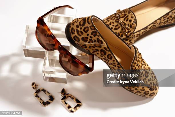 set of fashion collection with trendy fashion clothes for women loafer shoes in leopard print in beige and black colours, horn rimmed sunglasses, earrings on glass podium  on white color background. - leopardenfell stock-fotos und bilder