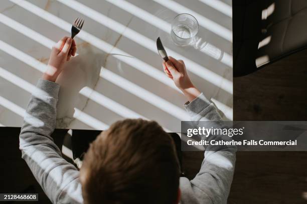 a child holds a knife and fork at a dining table - eating utensil stock pictures, royalty-free photos & images