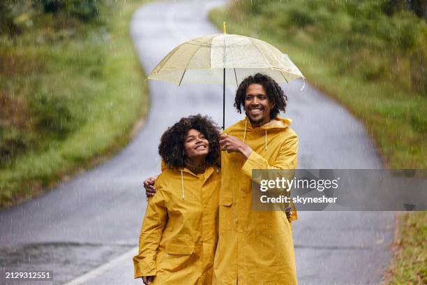 happy black couple walking embraced under umbrella on rainy day. - レインコート ストックフォトと画像