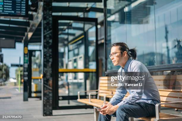 asian professional man sitting in a bus stop, holding a smartphone and looking into the distance - bus stop stock pictures, royalty-free photos & images