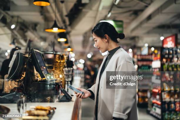 young asian woman making credit card mobile payment with her smartphone at a store, scan and pay a bill on a card machine making a quick and easy contactless payment at checkout counter. nfc financial technology, tap and go concept - digital counter stock pictures, royalty-free photos & images