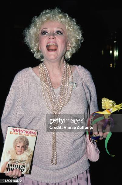 American comedian and actress Phyllis Diller, wearing a sweater, with a multi-strand beaded necklace, holding a copy of her book 'The Joys of Aging &...