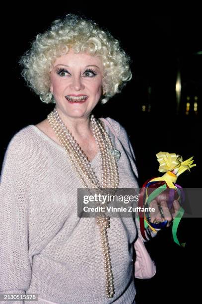 American comedian and actress Phyllis Diller, wearing a sweater, with a multi-strand beaded necklace, holding a red onion wrapped in ribbon in her...