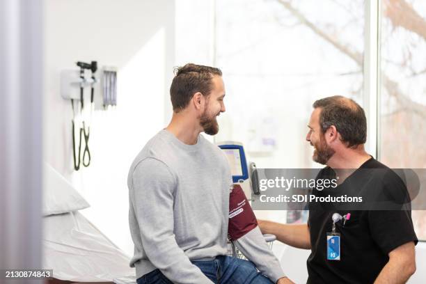 male doctor encourages young man about blood pressure reading - examination table stock pictures, royalty-free photos & images