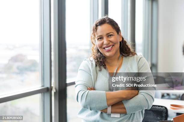 cheerful female teacher stands with arms crossed in classroom - mid adult stock pictures, royalty-free photos & images