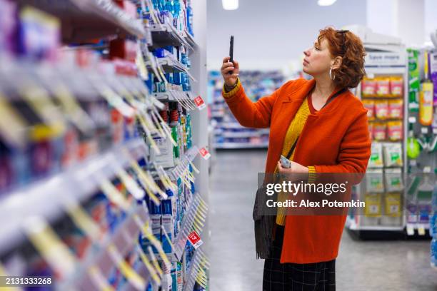 femme en cardigan orange utilisant un smartphone pour scanner l’étiquette de l’article dans la section des produits d’entretien ménager du supermarché - produit dentretien photos et images de collection