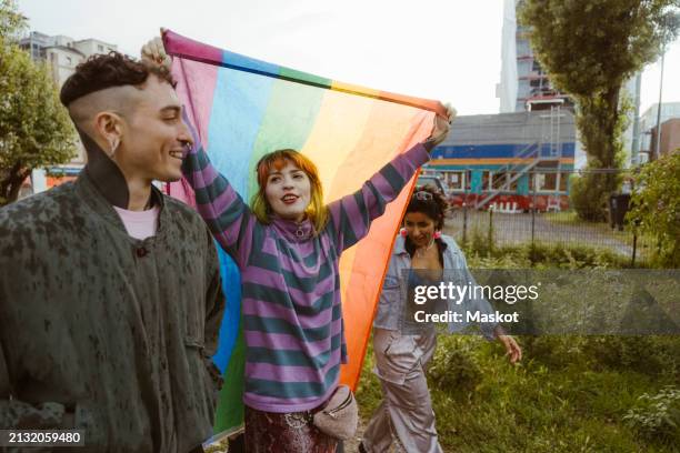 woman holding lgbt flag while walking with non-binary friends at park - berlin gay pride stock pictures, royalty-free photos & images