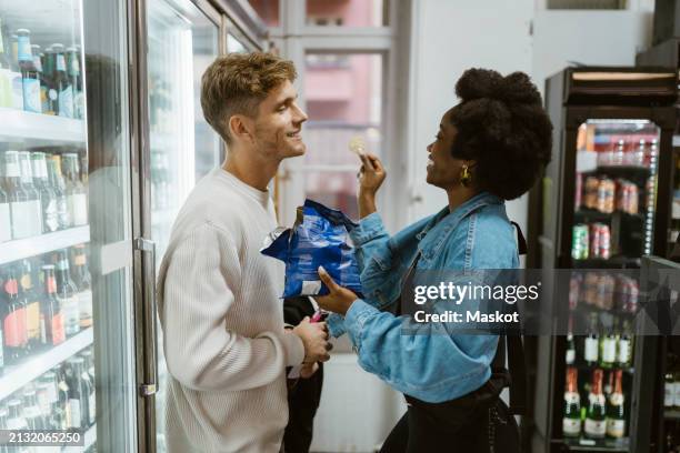 side view of girlfriend feeding chips to boyfriend while standing at aisle at supermarket - snack aisle stock pictures, royalty-free photos & images
