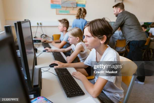 concentrated male pupil using computer while sitting with students at desk in computer classroom - computer lab kids stock pictures, royalty-free photos & images