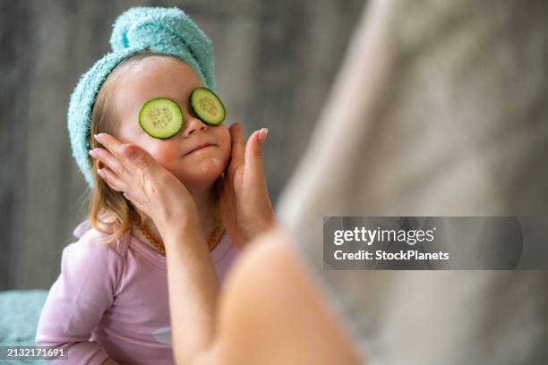 a little girl with cucumber slices and a towel on her head enjoys a spa day at home - mom and daughter pamper stock pictures, royalty-free photos & images