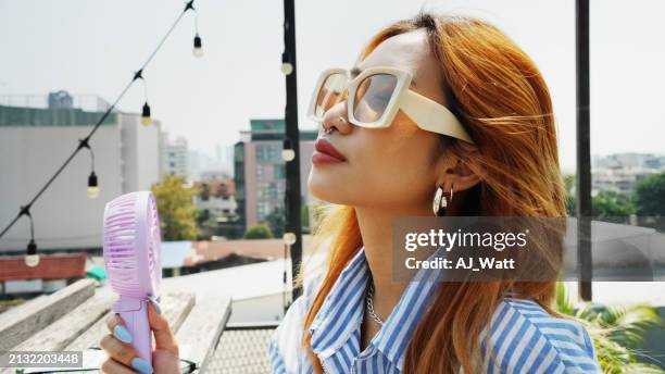 asian young woman holding portable electric fan near her face at rooftop cafe - hand fan stock pictures, royalty-free photos & images