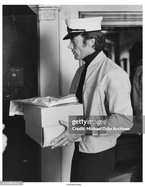 American actor and film director Clint Eastwood, wearing a peaked cap while carrying a box in a publicity still for 'The Eiger Sanction', 1975....