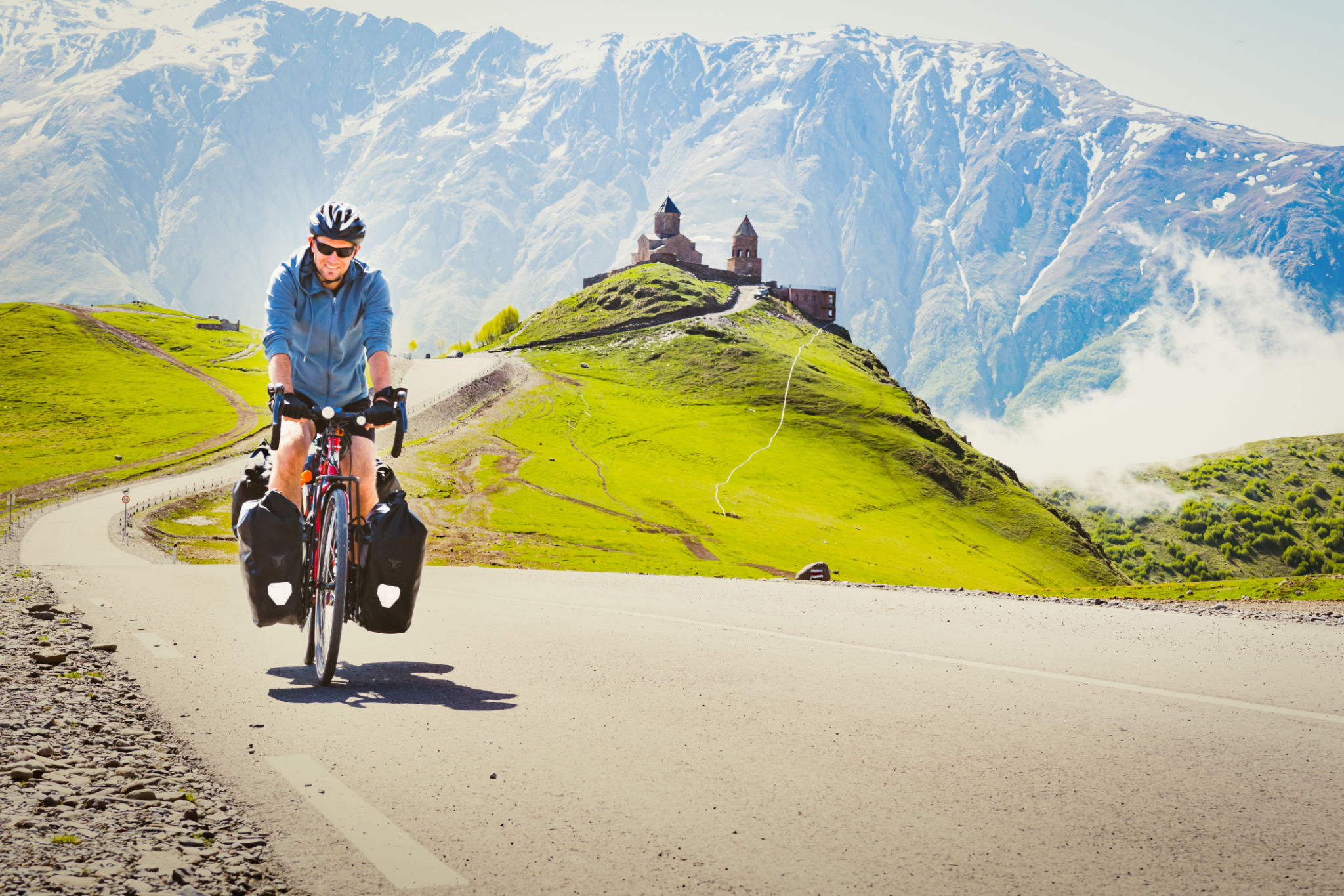 Male cyclist on touring bicycle adventure cycle visits Gergeti trinity church with mountains background. Cycling holidays and travel around caucasus mountain range Male cyclist on touring bicycle adventure cycle visits Gergeti trinity church with mountains background. Cycling holidays and travel around caucasus mountain range