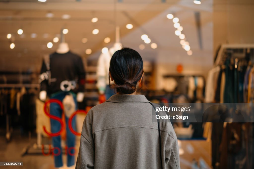Rear view of young Asian woman standing outside a boutique looking at clothings through shop window while shopping in shopping mall. Window shopping. Sale season. Seasonal shopping and consumerism concept