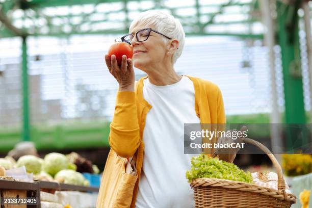 portrait of a senior woman holding basket full of fresh groceries and smelling organic tomato - odorat photos et images de collection