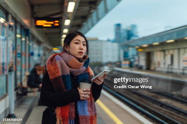 young asian woman checking time via smartphone while waiting for the train on railway platform - railway station platform stock pictures, royalty-free photos & images