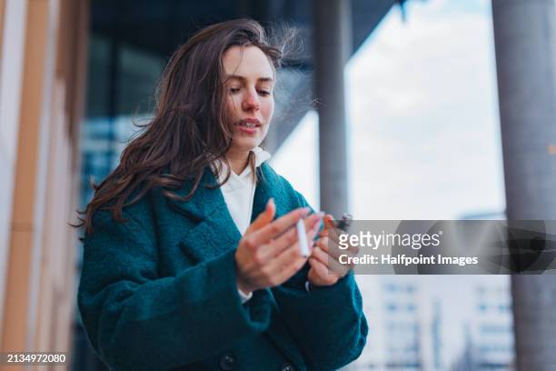 portrait of beautiful young woman smoking outdoors. standing on city street, holding cigarette and fire lighter. fashionable gen z woman with cigarette in mouth. - green lighter stock pictures, royalty-free photos & images