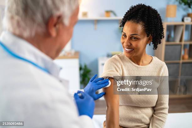 senior caucasian male doctor giving an flu vaccine to a female patient african-american ethnicity - flu vaccine stock pictures, royalty-free photos & images