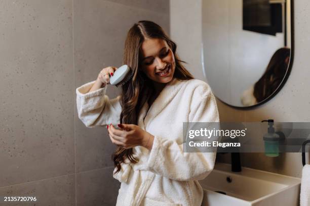 donna che pettina i suoi bei capelli con la spazzola mentre si trova vicino allo specchio in bagno - cura dei capelli foto e immagini stock