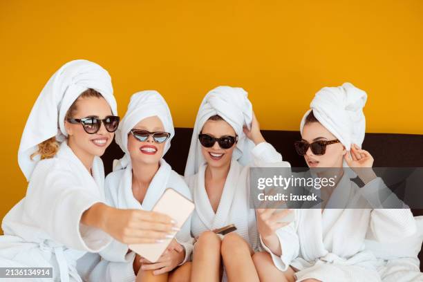 female friends wearing white bathrobes and towels on heads sitting on bed and using smart phones - hotel room white bed stock pictures, royalty-free photos & images