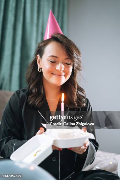 smiling young brunette woman in black pajamas with birthday cake in hands making wish sitting on bed at home - birthday candle number stock pictures, royalty-free photos & images