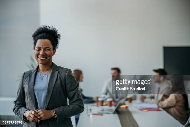 confident businesswoman standing tall, exuding leadership amidst a bustling office meeting - exuding stock pictures, royalty-free photos & images