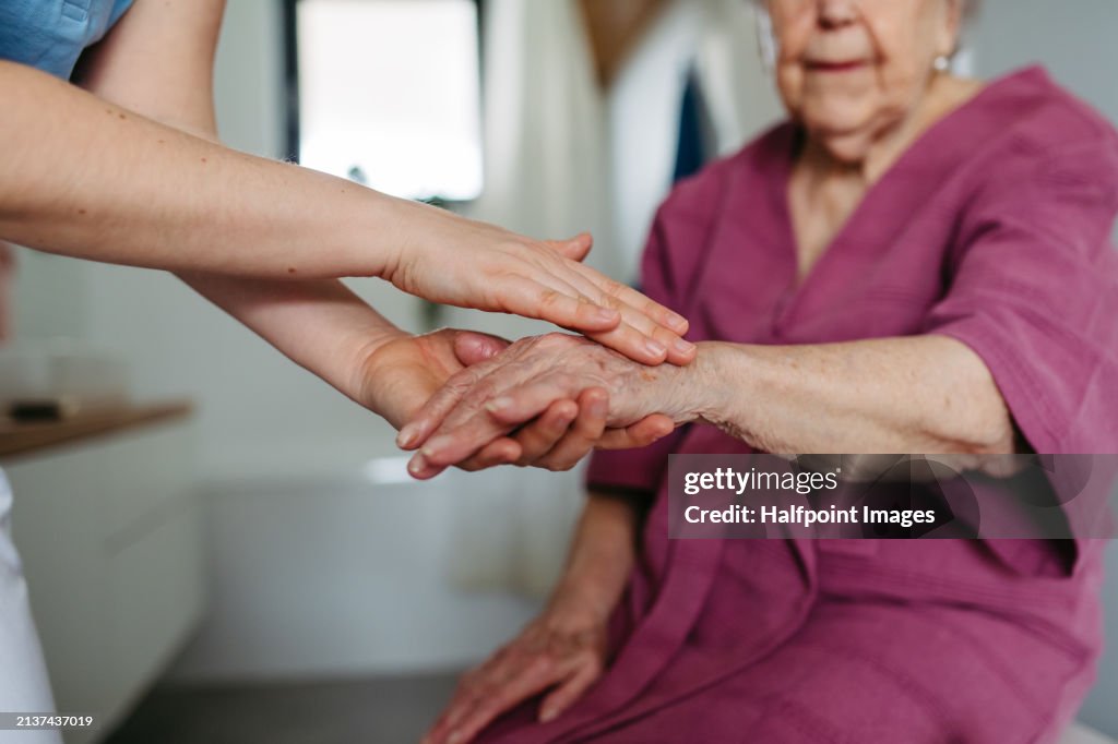 Home caregiver helping elderly woman with skincare in bathroom, applying moisturise lotion on hands, skin. Senior woman at home with nurse, close up.